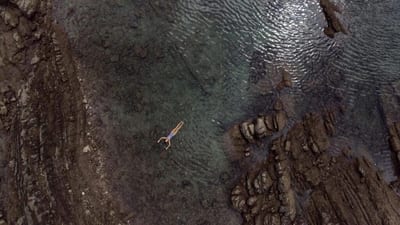 Aerial view of a person floating on their back in a clear natural tide pool surrounded by dark rocky formations on the Nicoya Peninsula, Costa Rica.