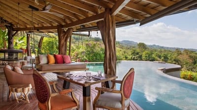 Open-air restaurant terrace with a curved wooden roof and a hanging daybed beside the infinity pool overlooking the rainforest at Origins Lodge in Bijagua, Costa Rica