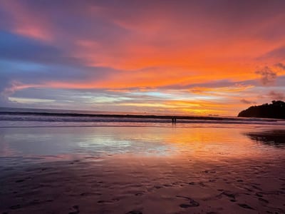 A vivid orange, pink, and purple sunset blazes over the Pacific and mirrors on the glassy wet sand, with two distant figures at the waterline and a forested headland on Costa Rica's Pacific coast.