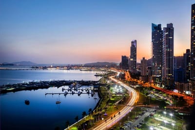 Twilight view over Panama City with lit high-rise towers, the curving Cinta Costera highway, and a marina full of boats in the bay