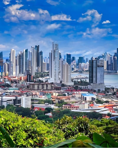 The high-rise skyline of Panama City rising above a green hillside, with the Pacific bay visible to the right under a blue sky.