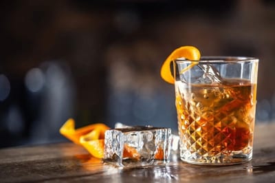 Amber Panamanian rum old fashioned in a faceted glass garnished with an orange peel twist beside a large ice cube on a rustic wooden bar