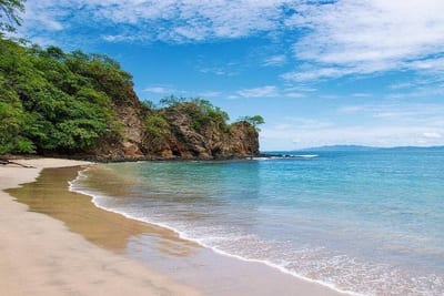 A quiet curve of pale-tan sand at Playa Penca meets calm, clear turquoise water that looks like a swimming pool, with a green-topped rocky headland to the left and distant land on the horizon, Guanacaste, Costa Rica.