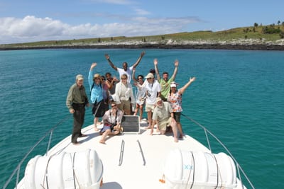 A group of travelers waving from the bow of a private boat on turquoise water off a green island in the Galápagos, Ecuador