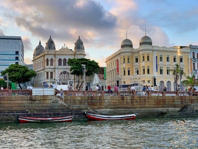 The colonial waterfront of Recife Antigo at sunset with fishing boats on the water and historic golden-lit buildings, Pernambuco, Brazil.