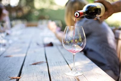 Red wine is poured from a bottle into a large stemmed glass on a weathered rustic wooden table, with people seated along the table in golden sunlight at a Mendoza vineyard, Argentina.