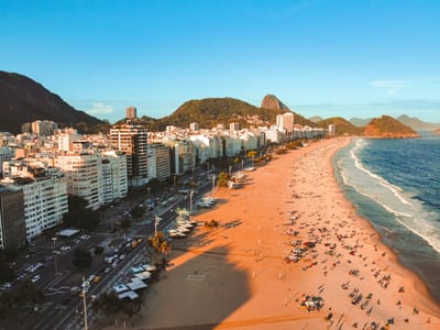 Aerial view of Copacabana beach at golden hour with its famous wave-patterned promenade curving along the Atlantic, Sugarloaf and the green hills of Rio de Janeiro in the distance, Brazil.