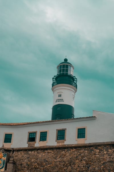 The green-and-white Farol da Barra lighthouse standing above its colonial fort under a cloudy sky in Salvador, Bahia, Brazil.