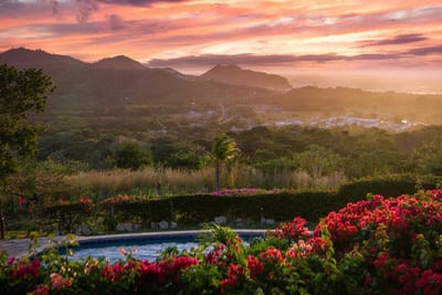 Hilltop view over forested hills and the Pacific at San Juan del Sur at sunset, framed by pink bougainvillea and a pool in Nicaragua