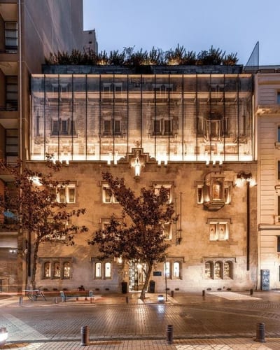 A restored heritage stone building with a glass-walled upper level and rooftop garden glows with warm light at twilight, framed by street trees, at a boutique hotel in central Santiago, Chile.