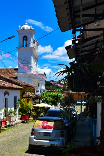 A cobblestone street in the colonial town of Suchitoto, El Salvador, leading toward the white bell tower of the Santa Lucía church, hung with papel-picado banners and lined by terracotta-roofed houses.