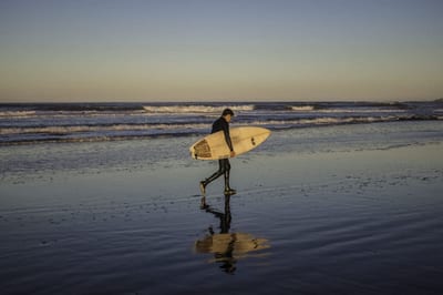Surfer in a wetsuit carrying a board along a wet-sand beach at sunset in Costa Rica