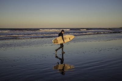 A surfer in a black wetsuit carries a cream surfboard across glassy, reflective wet sand at golden hour as small waves break behind on Costa Rica's Pacific coast.