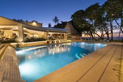 Glowing beachfront pool at dusk beside the open-air thatched bar at Tamarindo Diria Beach Resort, with palms silhouetted against the sunset over the Pacific in Guanacaste, Costa Rica