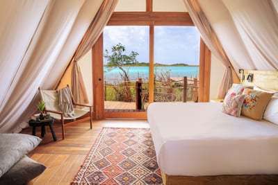The canvas-roofed interior of a luxury tented suite with a white bed and kilim rug opening through glass doors onto a deck above the turquoise Bacalar lagoon in Mexico.