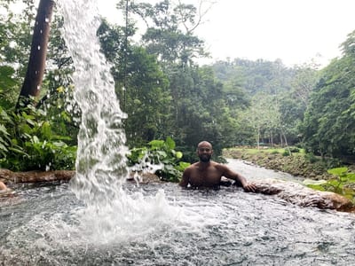 Man soaking in a jungle hot-spring pool beneath a cascading waterfall at Club Rio, The Springs Resort, beside the river in Arenal, Costa Rica