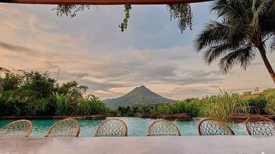 Arenal Volcano framed by palms and greenery across the infinity pool from the main pool bar at The Springs Resort &amp; Spa, with rattan bar stools in the foreground, Costa Rica