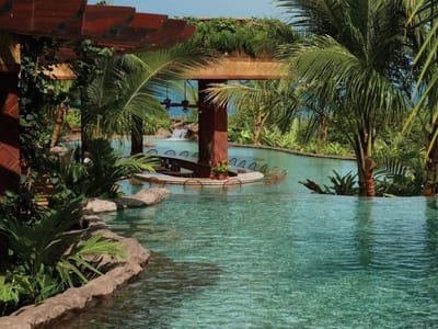 Swim-up pool bar with submerged stools under a wooden pergola, framed by palms and a waterfall, at The Springs Resort hot springs in Arenal, Costa Rica