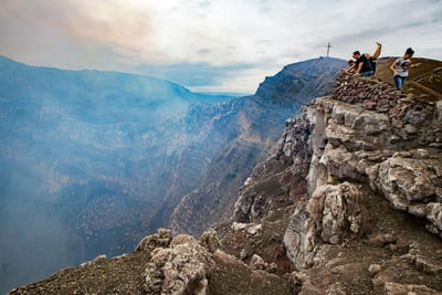A small group of tourists stands at a clifftop viewpoint beside a summit cross, gazing into the vast smoking crater of the active Masaya Volcano as volcanic gas rises from the depths, near Masaya, Nicaragua.