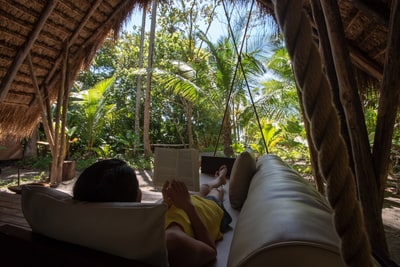 A traveler in yellow reclining on a daybed reads a book under a thatched-roof bungalow framed by palms and jungle at Isla Palenque, Panama