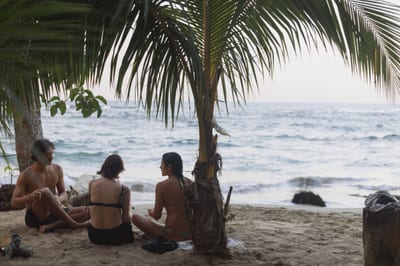 Three travelers in swimwear sitting and talking on the sand under a palm tree on a beach in Puerto Viejo, Costa Rica, with gentle waves behind them.