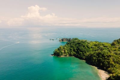 Aerial view of a jungle-covered peninsula reaching into the Pacific past rocky sea stacks, with a hidden sand cove, vivid turquoise-to-blue water, and a small boat's wake on Costa Rica's central Pacific coast near Manuel Antonio.