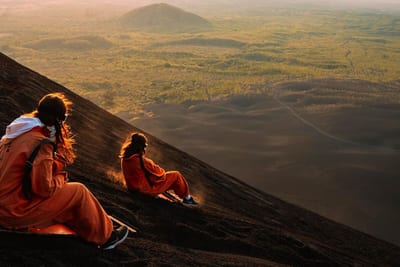 Two adventurers in orange jumpsuits sled down the black volcanic ash slope of Cerro Negro volcano above green plains at golden hour near León, Nicaragua.