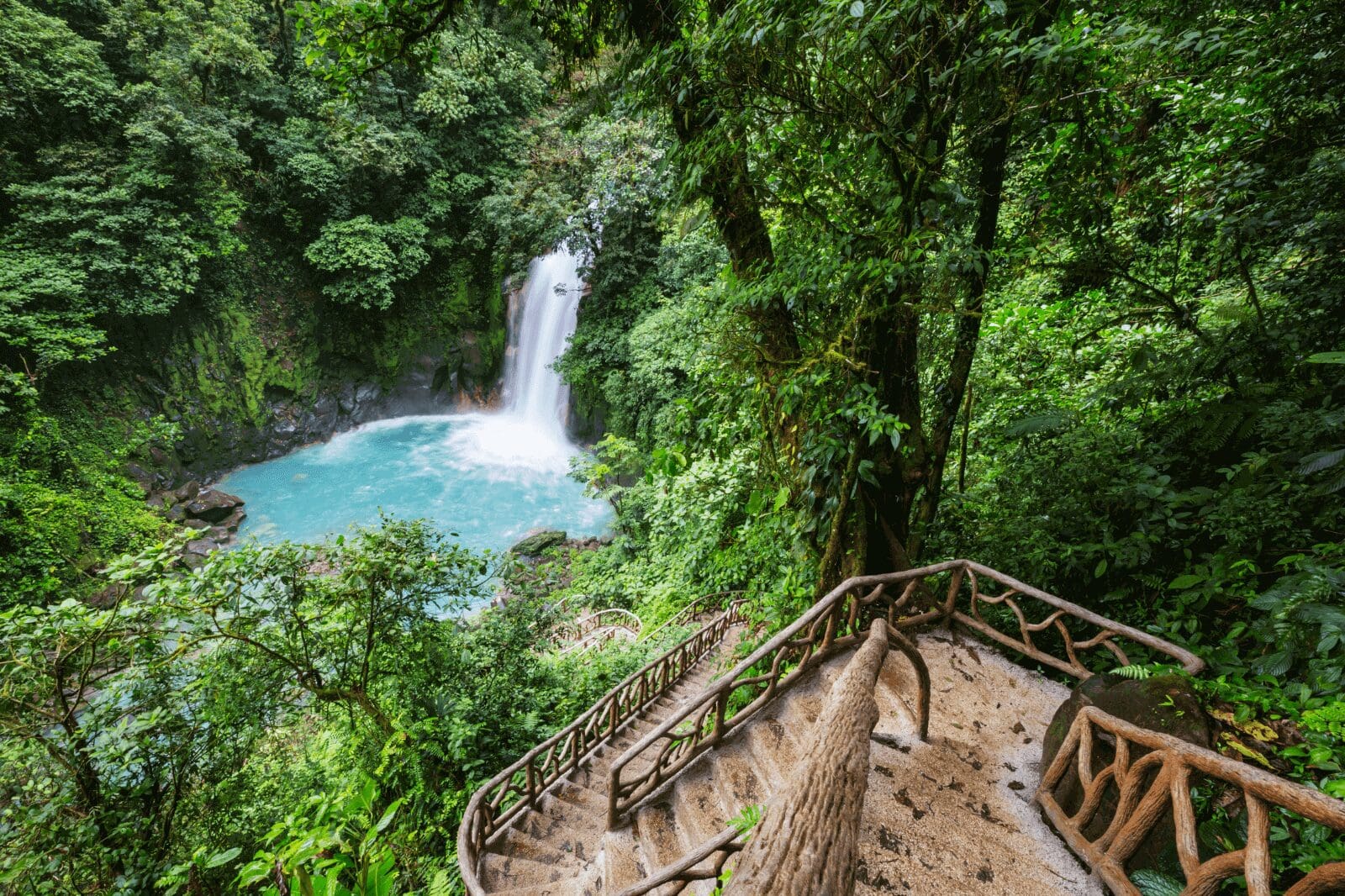 View of a jungle waterfall in Costa Rica seen from a lodge balcony, with surrounding lush greenery and a visible trail leading toward the falls.