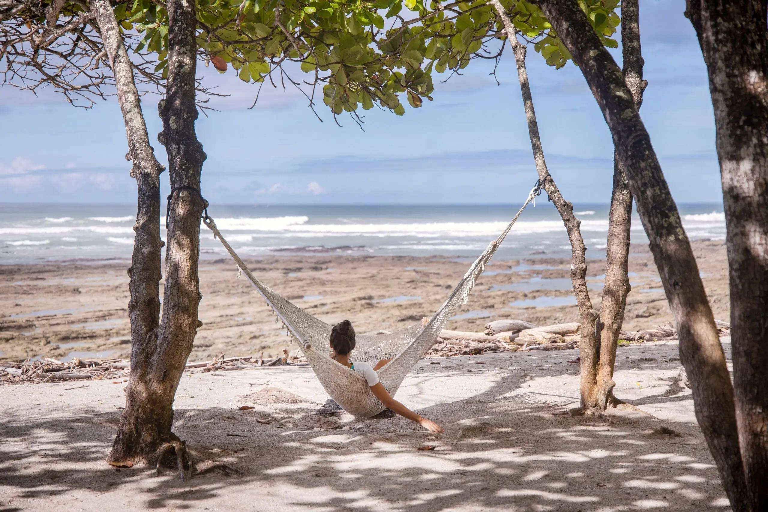 A person relaxes in a hammock tied between two trees on a sandy beach, facing the ocean. The tide is low, revealing rocky patches along the shore. Green leaves overhead provide dappled shade, while gentle waves roll in the distance under a partly cloudy sky.