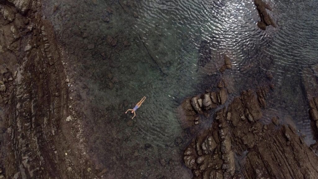 A person floats on their back in a shallow natural tide pool surrounded by rocky formations and clear water.
