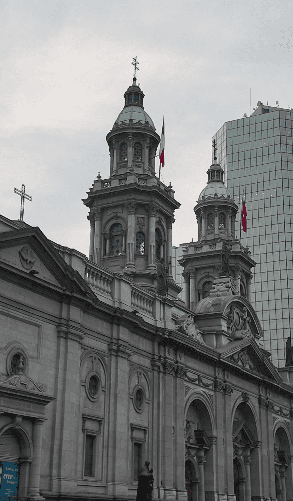 A historic cathedral with ornate twin towers topped by domes and crosses, each flying the Chilean flag. The stone façade features arched windows and intricate carvings, with a modern glass skyscraper rising in the background under a cloudy sky.