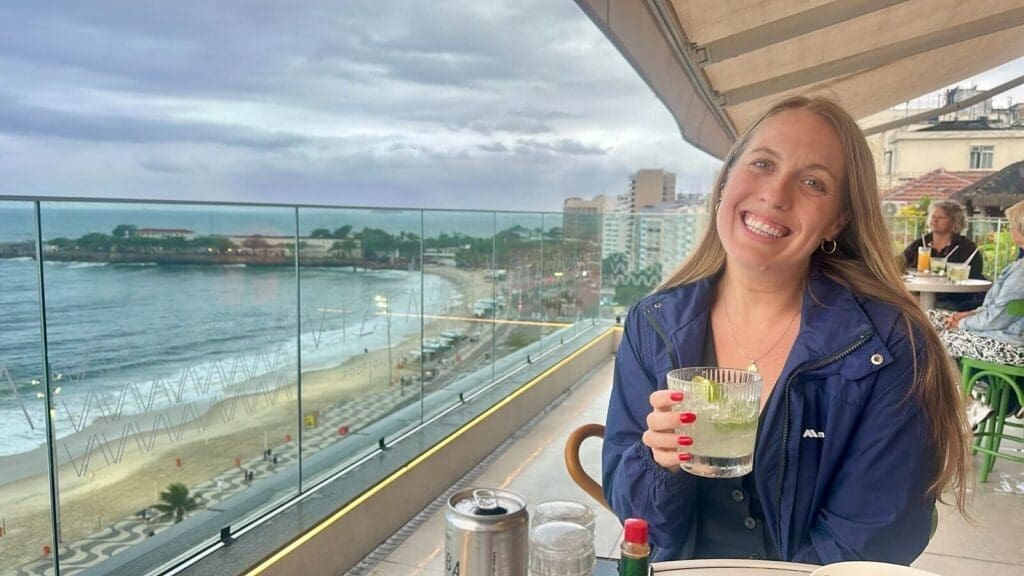 A woman, Kathleen Hall, smiles while sitting at an outdoor restaurant overlooking a beachside cityscape. She is holding a drink with ice and lime, wearing a blue jacket, and seated at a table. The ocean, boardwalk, and distant buildings are visible under a cloudy sky.