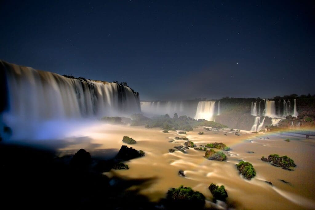 A breathtaking nighttime view of a wide, multi-tiered waterfall cascading over rocky cliffs. The water appears silky under long exposure, reflecting soft moonlight. Mist rises from the falls, and a faint rainbow glows near the lower right, while patches of green moss dot the smooth, flowing riverbed beneath a starry sky.