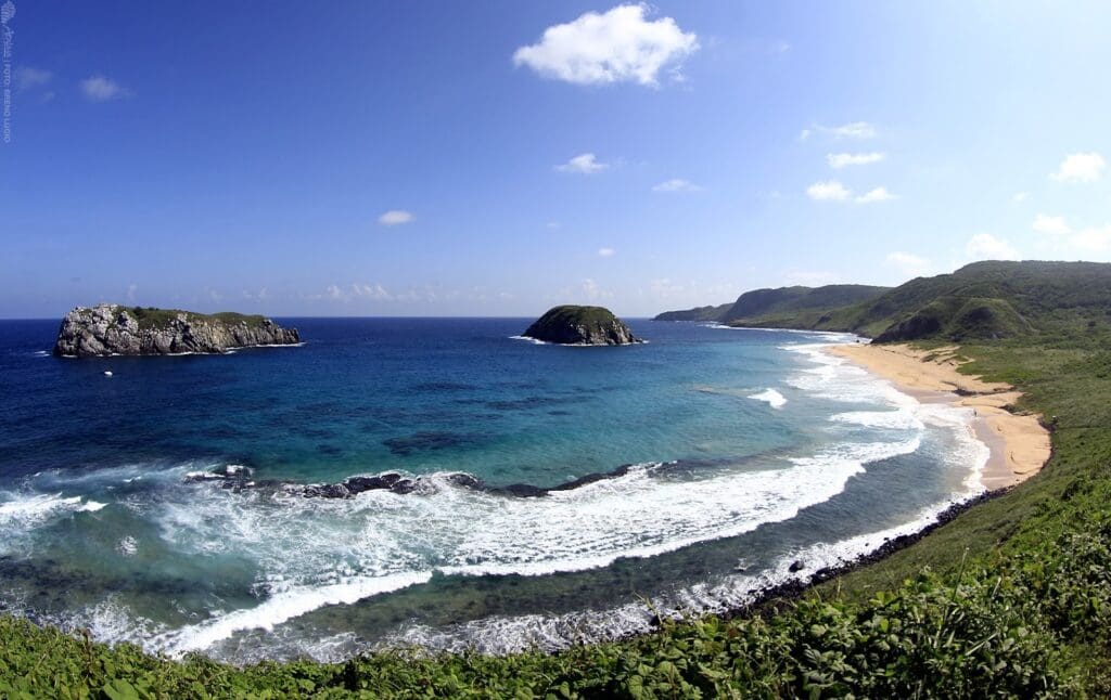A panoramic view of a pristine coastal landscape with turquoise waves crashing onto a golden sandy beach. Two small rocky islands rise from the deep blue ocean near the shore, surrounded by lush green hills under a bright, clear sky with scattered white clouds.