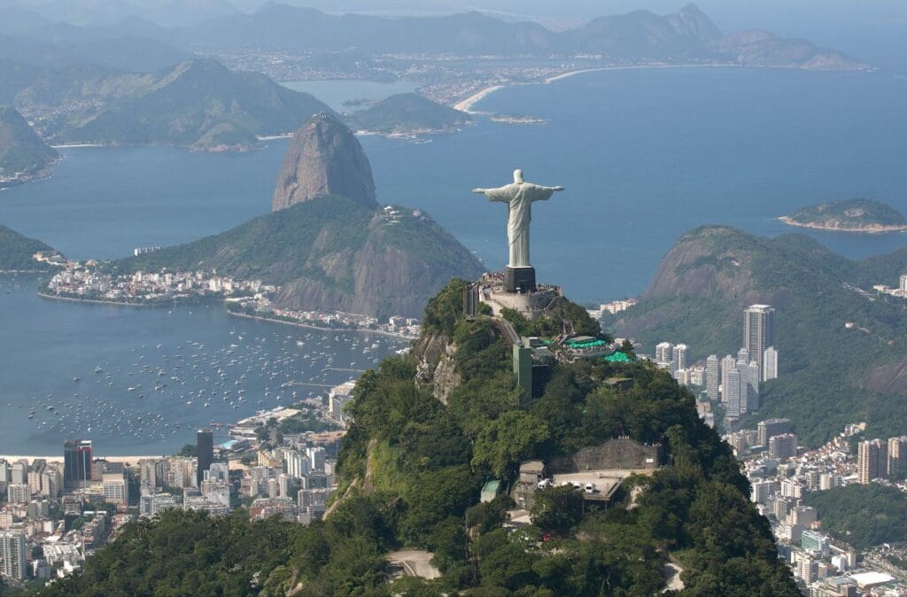 Aerial view of the Christ the Redeemer statue atop Corcovado Mountain, overlooking Rio de Janeiro, Brazil. The vast cityscape spreads below with Sugarloaf Mountain rising near the coastline, boats dotting the blue bay, and lush green hills surrounding the area under a clear sky.