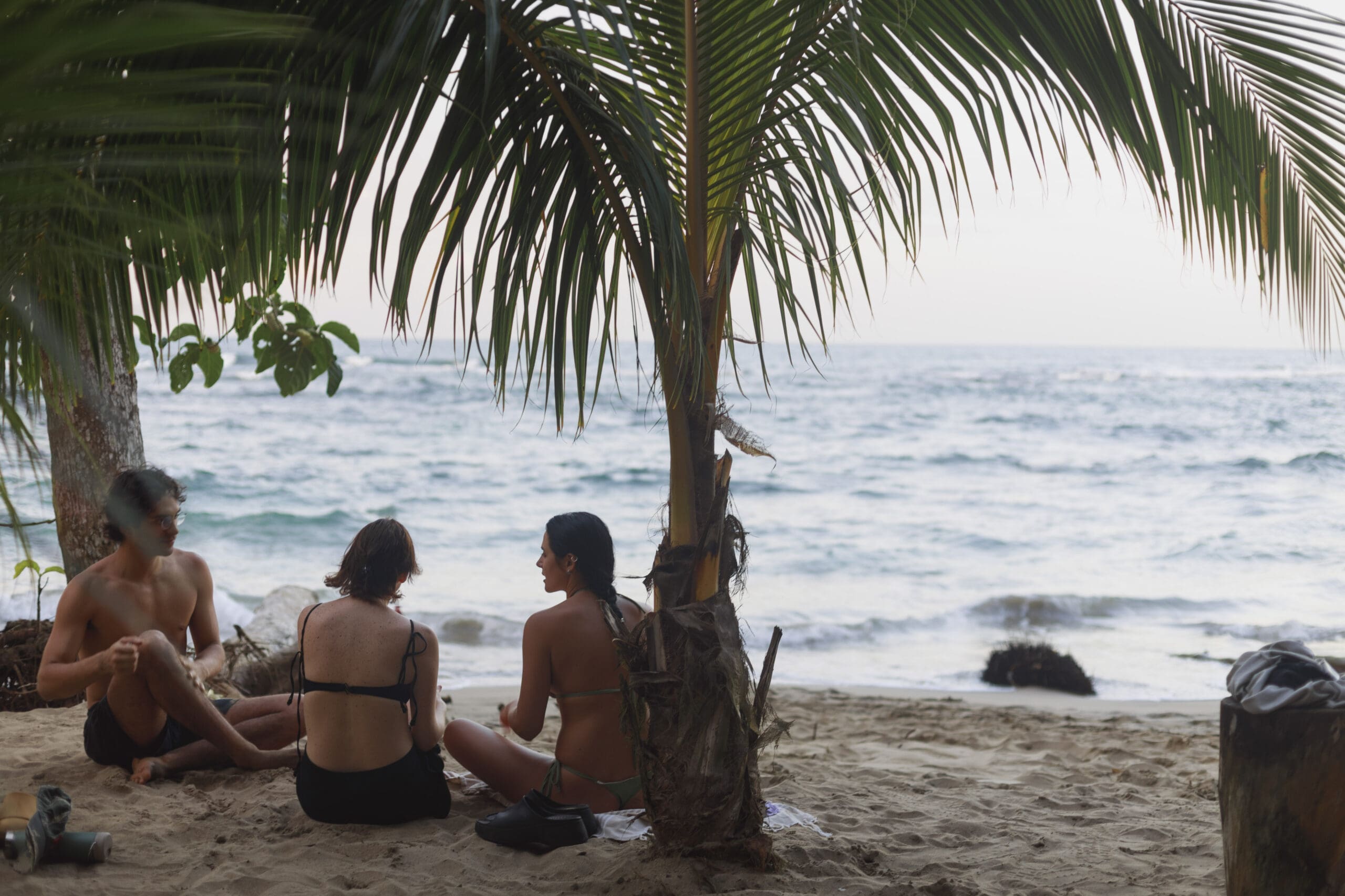 Group of travelers sitting under a palm tree on a sandy beach in Puerto Viejo, Costa Rica, with waves and the Caribbean Sea in the background.