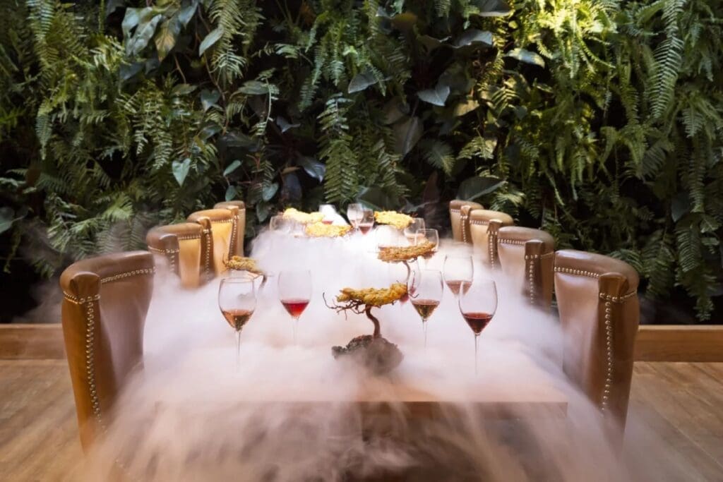 Elegant dining table with wine glasses and artistic food presentation surrounded by theatrical fog, set against a lush green plant wall at Elcielo Restaurant Medellin.
