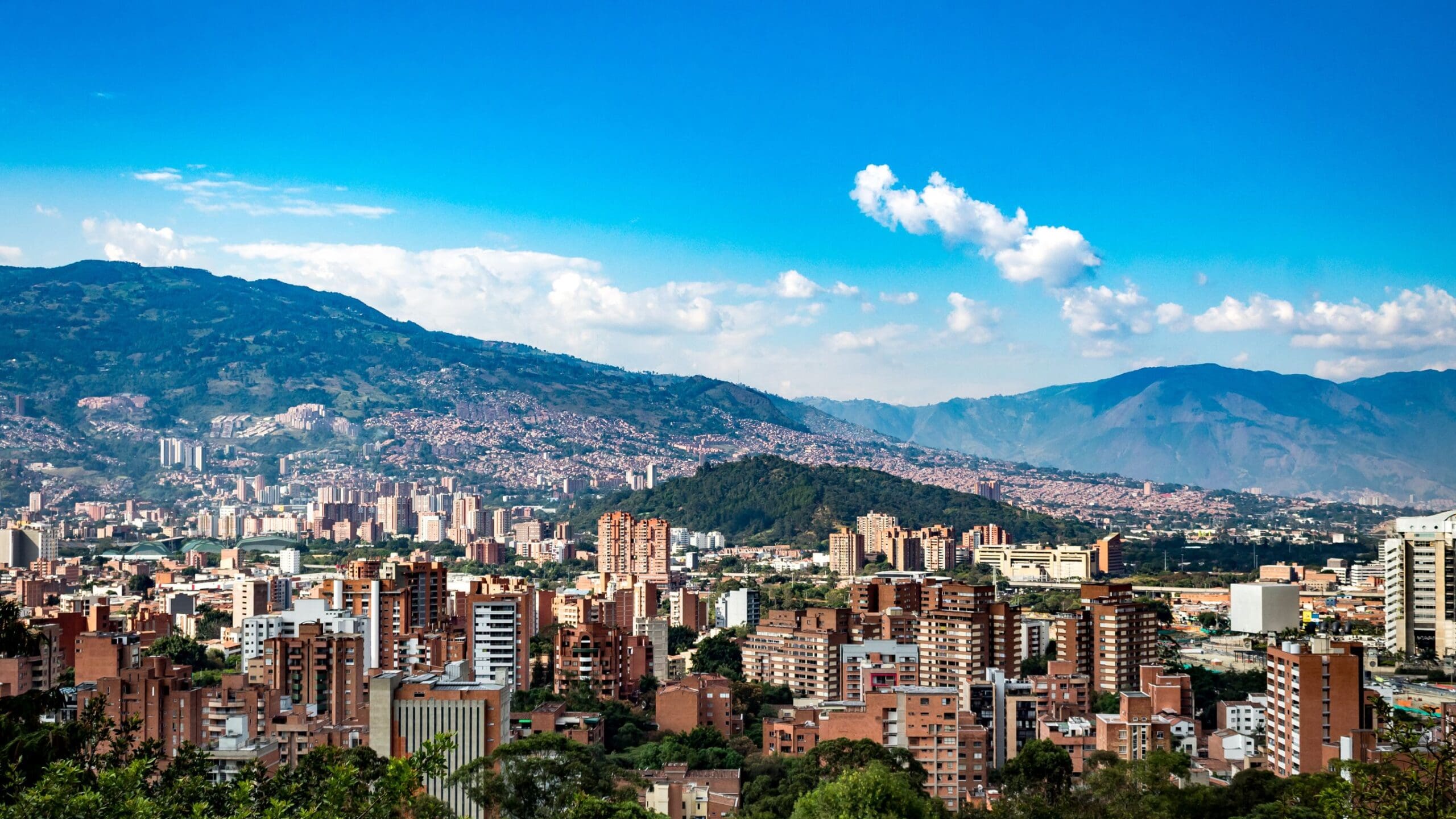Panoramic view of Medellín with its high-rise buildings, lush green hills, and mountains under a bright blue sky.